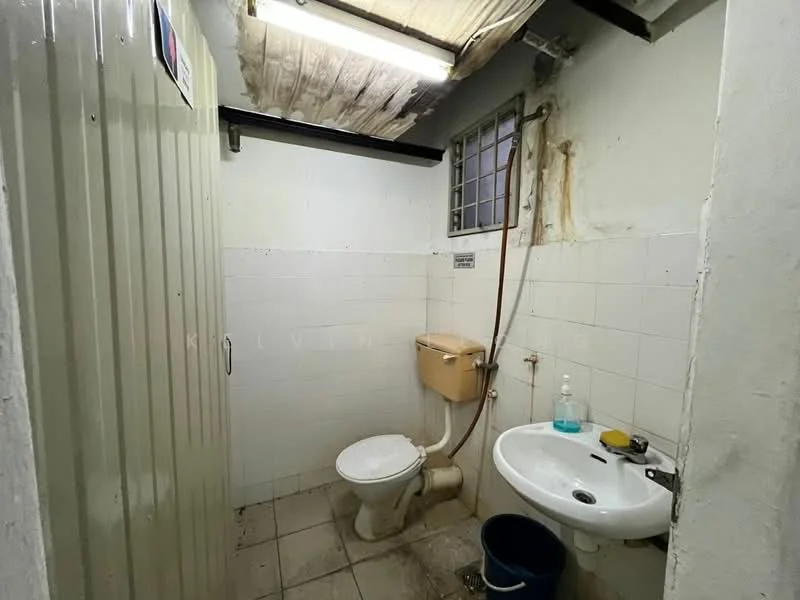 Bathroom features: white tiled walls, white ceramic floor tiles, one-piece toilet, wall-mounted sink, chrome faucet, shower hose, small window, plastic bucket, exposed plumbing, fluorescent ceiling light, metal partition, water bottle on sink.