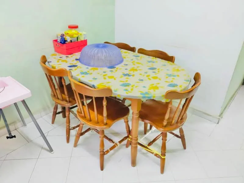 Dining features: oval dining table with floral tablecloth, 6 wooden dining chairs, light green painted walls, white tiled flooring, plastic storage container on table, basket with food items on table.