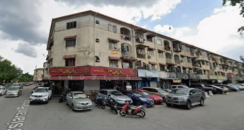 Exterior type: low-rise apartment block; features: multi-storey building, commercial units on ground floor, balconies on upper floors, exposed concrete facade, signage for businesses, parking lot in front, various parked vehicles, surrounding greenery; style: traditional.