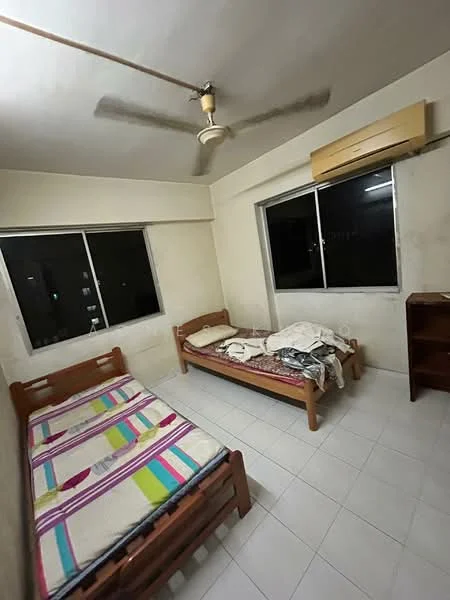Bedroom features: two single beds with wooden frames, bedding with colorful stripes, white tiled flooring, ceiling fan, wall-mounted air conditioning unit, large window with black frames, wooden bookshelf, plain white walls.