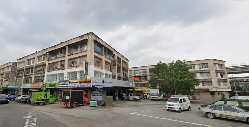 Exterior type: shophouse; features: 3-storey building, concrete facade, balconies with railings, ground floor retail spaces, parking area in front, multiple vehicle access, signage on storefronts, trees in landscaping, overcast sky; style: traditional.