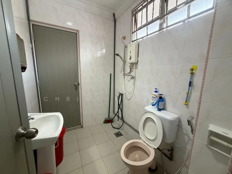 Bathroom features: white tiled walls, light grey floor tiles, single-sink vanity with cabinet, chrome basin mixer faucet, one-piece toilet, shower with wall-mounted shower head, water heater, cleaning supplies visible, window for natural light, door with handle.