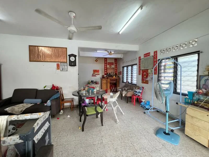 Living features: grey tiled flooring, white painted walls, ceiling fan, wall-mounted clock, large windows with grilles, wooden sideboard, round dining table, black sofa, plastic chairs in various colors, decorative wall hangings, floor fan.