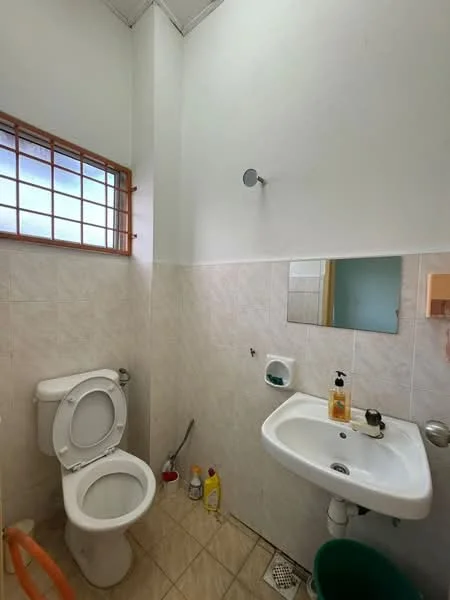 Bathroom features: one-piece toilet, single-sink vanity with wall-mounted faucet, framed mirror, tile cladding on walls, ceramic tile flooring, window with wooden grille, soap dispenser, toilet paper holder, cleaning supplies visible.