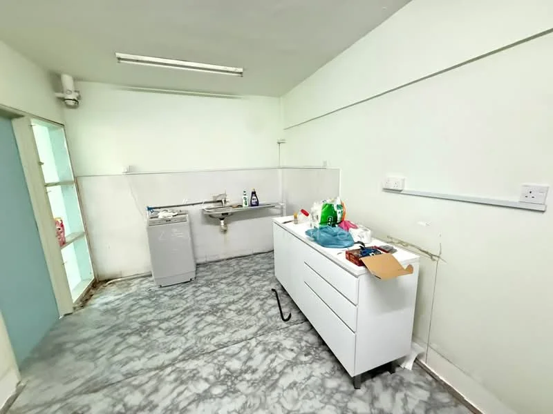 Kitchen features: white cabinetry with drawers, stainless-steel sink, chrome faucet, grey marble flooring, white walls, overhead fluorescent lighting, window with light blue frame, washing machine, countertop with clutter.