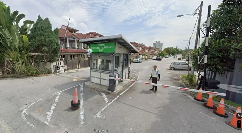 Exterior type: main entrance gate; features: guard house at entrance, barrier gate, traffic cones, paved road, surrounded by greenery, residential buildings in background; style: modern.