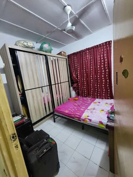 Bedroom features: single bed with pink bedding, wooden wardrobe with sliding doors, curtains with floral pattern in red, white tiled flooring, ceiling fan, decorative items on top of wardrobe, suitcase on the floor, wall with visible paint peeling.