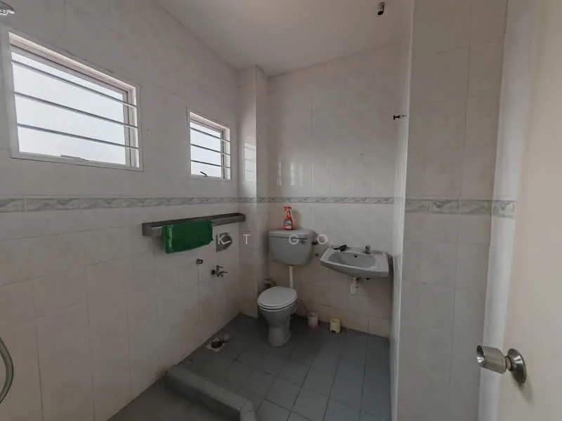 Bathroom features: white tiled walls, grey tiled flooring, one-piece toilet, wall-mounted sink, chrome faucet, small window for natural light, green towel hanging, toilet paper holder, soap dispenser, cleaning supplies on floor.