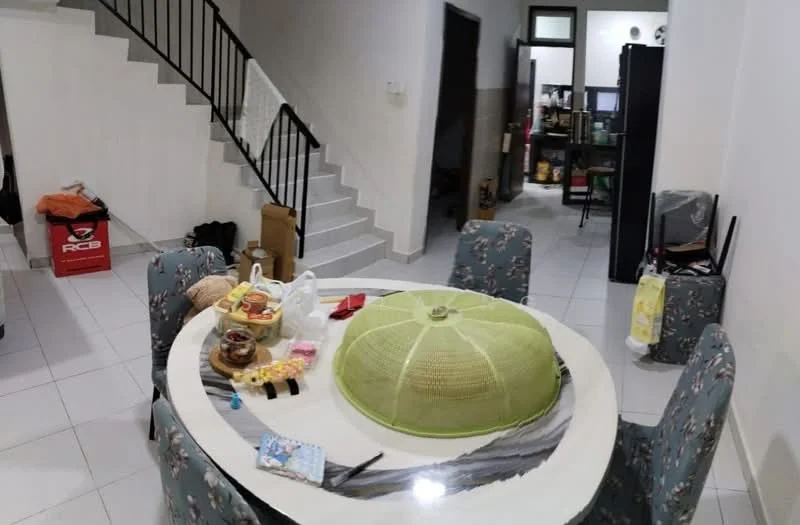 Dining features: round dining table, upholstered dining chairs with floral pattern, white walls, tile flooring, ceiling light fixture, decorative tablecloth, food items on table, green food cover, open space leading to staircase. Room features: staircase leading to upper floor, open kitchen area visible in background, black refrigerator, shelves with storage items.