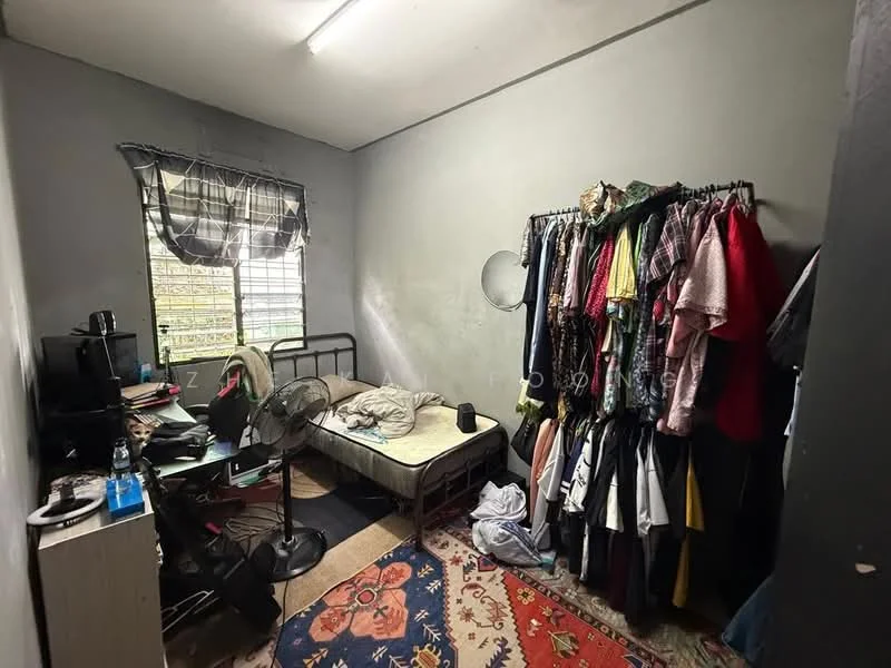 Bedroom features: grey painted walls, vinyl flooring, single bed, metal bed frame, clothes rack with various clothing, desk with computer monitor, desk lamp, ceiling light, window with sheer curtains, area rug with floral pattern, standing fan.