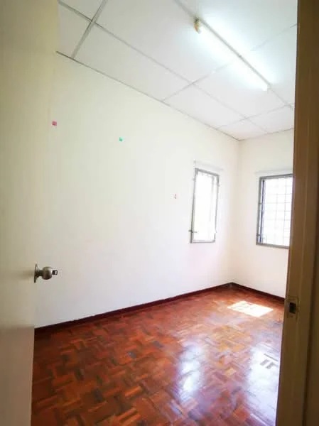 Room features: white painted walls, parquet wood flooring, two small windows with metal grills, fluorescent ceiling light, plain ceiling, door with handle.