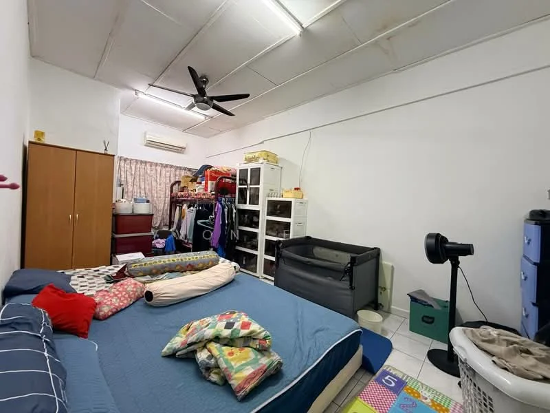 Bedroom features: queen-size bed with blue bedding, colorful quilt, wooden wardrobe with sliding doors, white shelving unit, clothes hanging on rack, floor fan, ceiling fan, white walls, tile flooring, laundry basket, playmat with numbers, crib, curtains, air conditioning unit.