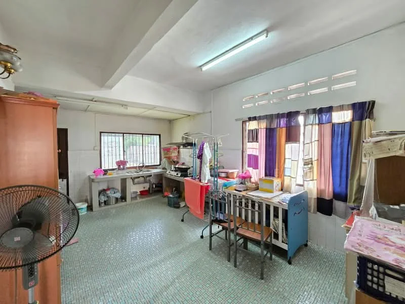 Room features: green mosaic tile flooring, white painted walls, ceiling fan, window with sheer curtains, window with colored curtains, wooden cabinet, metal drying rack, folding table, plastic chair, sink with countertop, laundry area, open shelving, various household items on surfaces.