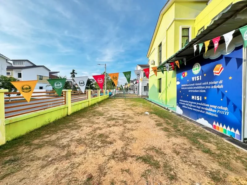 Exterior features: yellow painted facade, decorative flags along the pathway, manicured lawn, fenced perimeter, blue wall with text, pathway leading to entrance, surrounding residential buildings; style: modern.