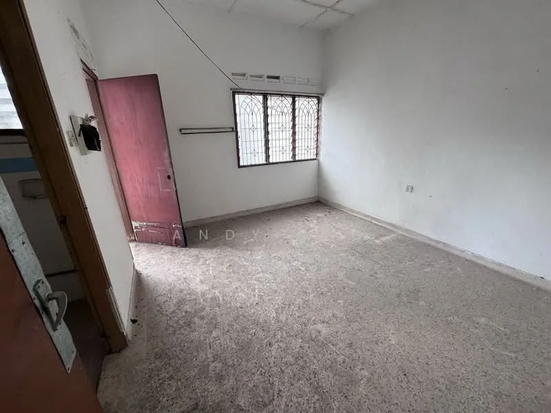 Room features: white painted walls, concrete flooring, single window with metal grille, wooden door, no visible furniture, ceiling with exposed beams, light fixture on ceiling, corner of room with potential for storage.