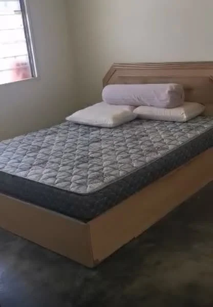 Bedroom features: queen-size bed, wooden bed frame, mattress with quilted cover, two pillows, concrete flooring, window with natural light, plain white walls; style: minimalist.
