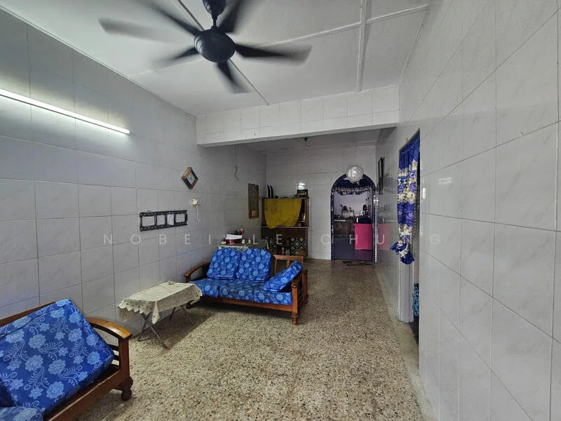 Living features: blue floral fabric sofa, wooden coffee table, ceiling fan, white tiled walls, grey speckled flooring, kitchen area visible in background, blue patterned curtains, wall-mounted light fixture.