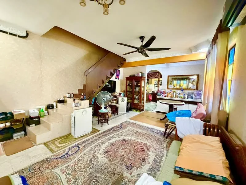 Living features: cream painted walls, exposed beams on ceiling, 5-blade ceiling fan, wall-mounted air conditioning unit, large area rug with intricate pattern, wooden sofa with cushions, wooden coffee table, two small wooden chairs, staircase leading to upper floor, decorative wall art, large window with sheer curtains, open layout to kitchen area; style: traditional.