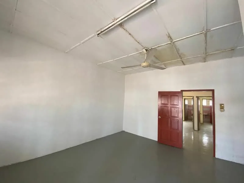 Room features: white painted walls, grey flooring, exposed ceiling with metal beams, ceiling fan, single red door, door frame with no visible trim, light fixture on ceiling.
