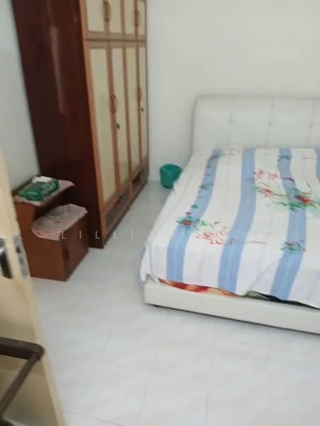 Bedroom features: queen-size bed with white leather headboard, floral patterned bedspread, white tiled flooring, wooden wardrobe with sliding doors, two wooden storage boxes, green plastic bucket, plain white walls, natural light from window.