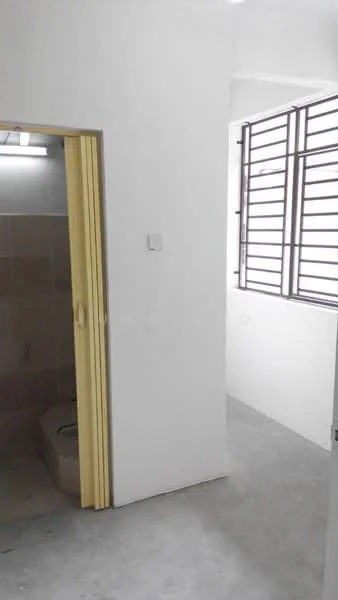Room features: white painted walls, concrete flooring, window with horizontal metal grilles, door with yellow frame, shower area with tiled walls, toilet bowl, light fixture on ceiling.