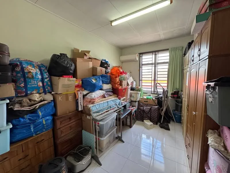 Room features: white painted walls, glossy tile flooring, window with blinds, air conditioning unit, wooden wardrobe, plastic storage boxes, stacked cardboard boxes, wooden chest of drawers, metal shelving unit, plastic containers, folding table, various items cluttered, green curtains, overhead fluorescent lighting.