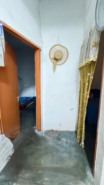 Room features: white painted walls, concrete flooring, wooden door, doorway leading to another room, hat hanging on wall, curtain with gold color, blue bedding visible in adjacent room.