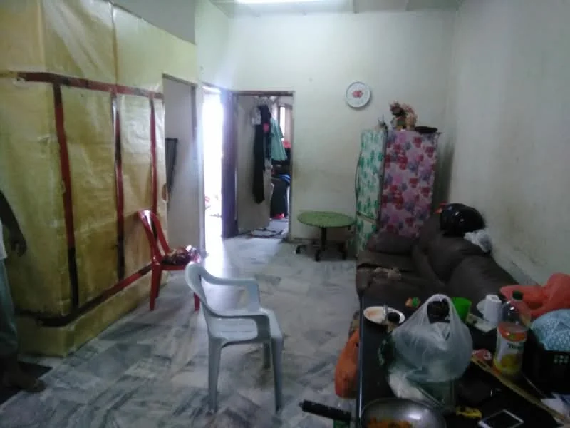 Living features: marble flooring, brown leather sofa, plastic chair, round green table, wall with yellow covering, decorative wall clock, clothes hanging on door, kitchen utensils on table, open doorway, messy countertop.