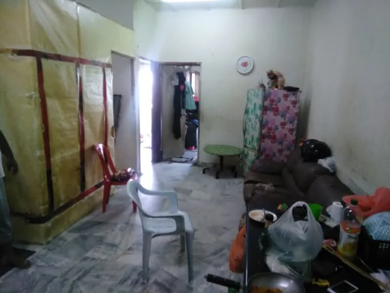 Living features: marble flooring, brown leather sofa, plastic chair, round green table, decorative floral cabinet, wall with cream paint, doorway leading to another room, visible clutter on table, clothes hanging in doorway, partition with yellow plastic covering.