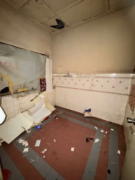 Room features: unfinished walls, dirty floor tiles, broken ceiling tiles, exposed electrical wiring, window with torn curtain, scattered debris on the floor, stained wall tiles, hanging clothes hanger.