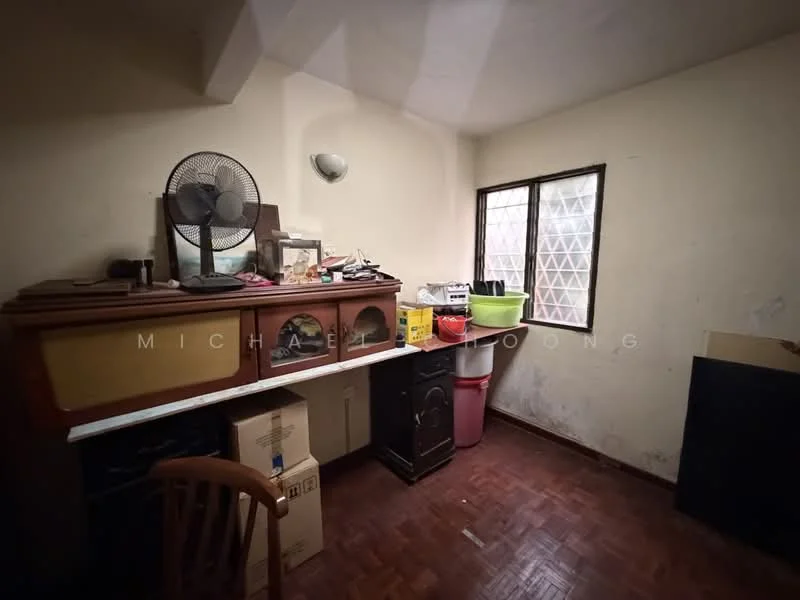 Room features: wooden flooring, white painted walls, single window with metal grille, ceiling light fixture, wooden sideboard, plastic trash bin, cardboard boxes, fan, table, various items on sideboard.