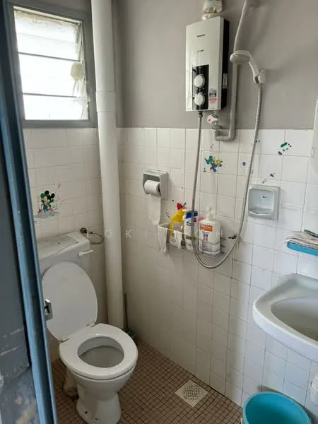 Bathroom features: white toilet, wall-mounted shower with water heater, sink with wall-mounted faucet, white tiled walls, light grey tiled flooring, toilet paper holder, shelf with toiletries, small drainage grate, window for natural light.