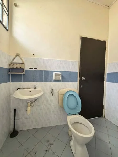 Bathroom features: white ceramic toilet, blue toilet seat, white sink with chrome faucet, grey tiled walls with blue accent tiles, grey floor tiles, wall-mounted soap dish, toilet paper holder, single mirror above sink, door with black finish, window for natural light.
