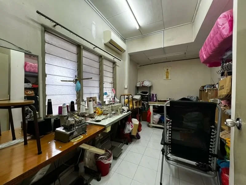 Room features: white painted walls, tile flooring, 1 wall-mounted split AC, multiple sewing machines, wooden table, plastic chairs, storage boxes, overhead lighting, large windows with sheer curtains, stacked fabric rolls, folding chair.