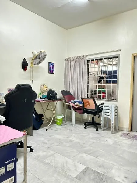 Room features: white painted walls, grey tiled flooring, window with metal grille, curtains, desk with chair, plastic stacking chairs, ceiling fan, clothes on chair, clothes on table, waste bin.