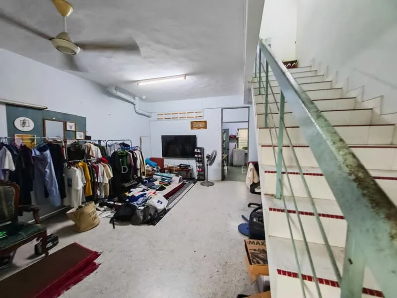 Room features: white painted walls, concrete flooring, ceiling fan, staircase with metal railing, large flat-screen TV, clothes hanging on racks, various boxes and items on the floor, area rug, light fixture on ceiling.