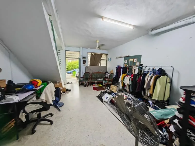Room features: white painted walls, concrete flooring, ceiling fan, overhead fluorescent lighting, large window with natural light, clothing rack with various garments, desk with office chair, folding table, boxes and miscellaneous items on the floor, decorative wall clock.