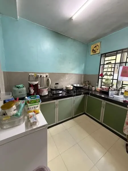 Kitchen features: light blue painted walls, grey tiled backsplash, green cabinetry, white countertop, stainless-steel sink, chrome faucet, built-in stove, microwave, kettle, rice cooker, various kitchen utensils, window with security bars, fluorescent ceiling light.