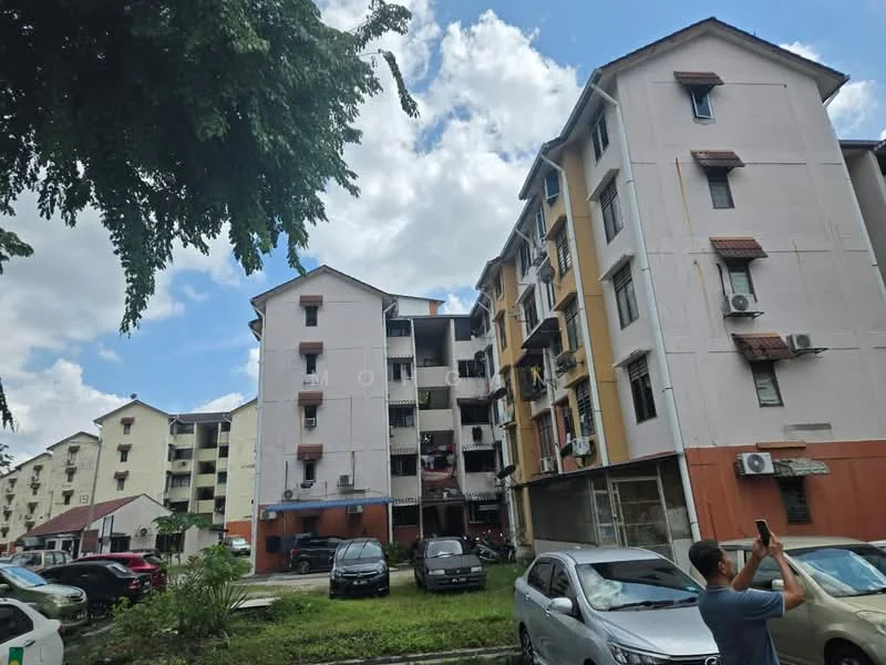 Exterior type: low-rise apartment block; features: 3-storey low-rise, painted facade in cream and orange, balconies with railings, visible air conditioning units, parking area with multiple cars, surrounded by greenery, cloudy sky, trees in foreground; style: modern.