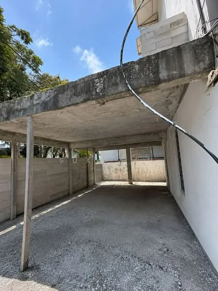 Exterior type: car porch; features: concrete flooring, exposed concrete ceiling, supporting wooden posts, surrounded by low concrete walls, open-air structure, direct sunlight, no visible doors or windows, gravel surface; style: minimalist.