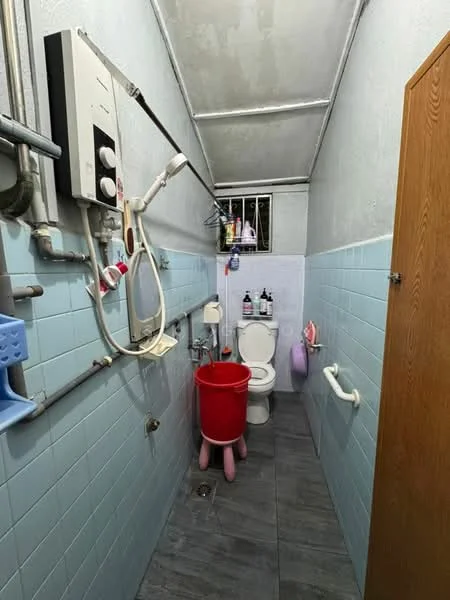 Bathroom features: toilet, shower with handheld showerhead, wall-mounted water heater, blue tiled walls, grey tile flooring, grab bars, window with security bars, wooden cabinet, red bucket, cleaning supplies on shelves.