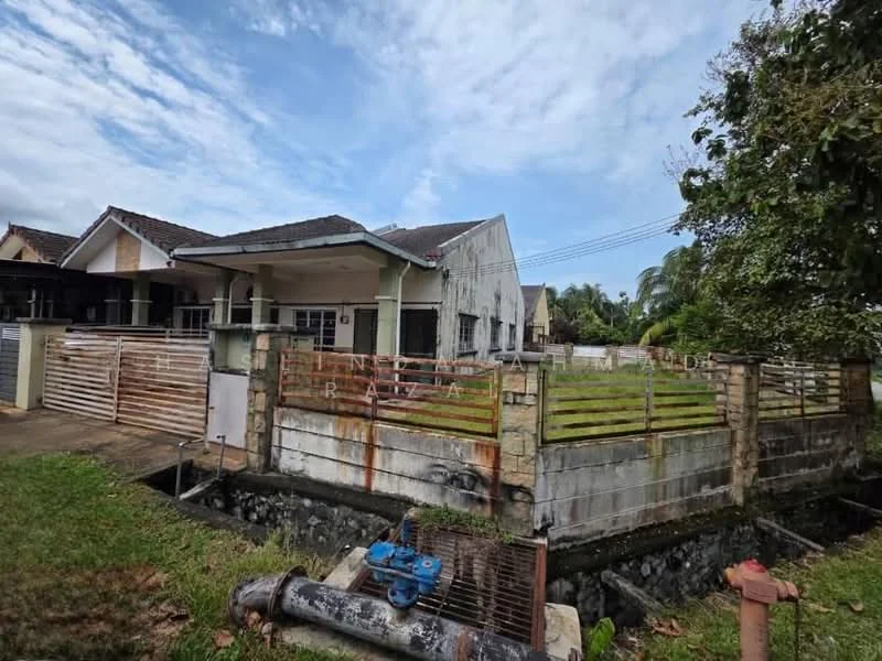 Exterior type: semi-detached house; features: weathered facade, single-storey structure, metal fencing, grassy yard, concrete pathway, visible drainage system, surrounding trees, partially cloudy sky; style: traditional.