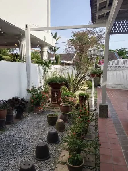 Exterior features: landscaped garden with various plants, stone-paved walkway, flower pots, decorative lanterns, white painted fence, gravel ground, paved pathway, mature trees, outdoor seating area; style: tropical-modern.