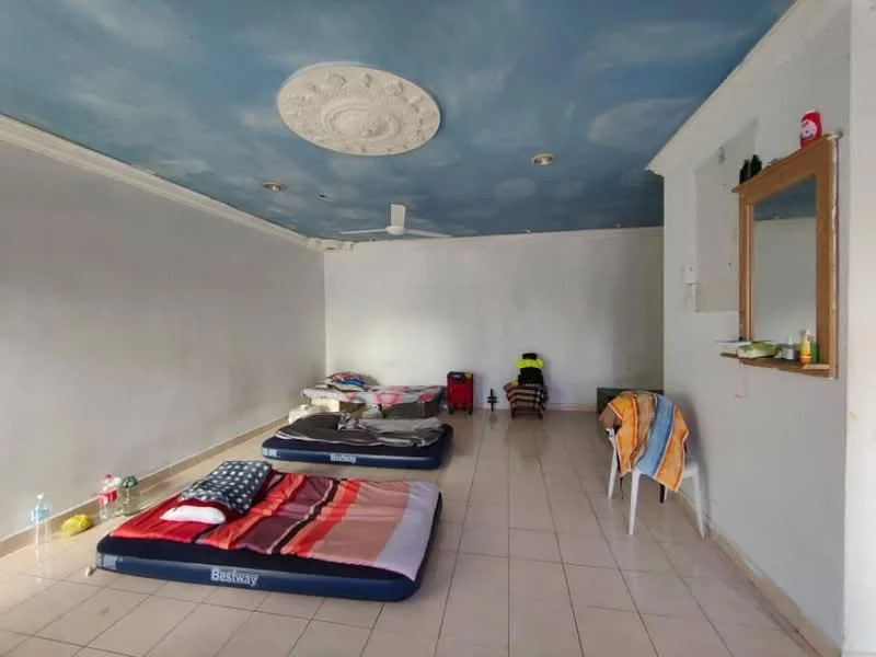 Room features: cream painted walls, blue painted ceiling with decorative plaster, tile flooring, 3 mattresses on the floor, 1 wall-mounted mirror, 1 ceiling fan, 1 small shelf with items, 2 bottles of water, 1 towel on a chair, 1 chair, 1 bag, 1 small table; style: minimalist.