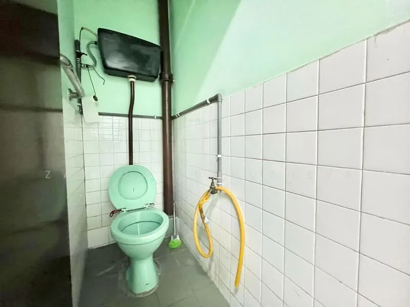 Bathroom features: green toilet, white tiled walls, grey tiled flooring, bidet spray, wall-mounted cistern, exposed plumbing, single wall-mounted light fixture.