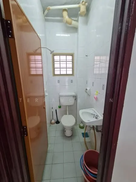 Bathroom features: white tiled walls, grey tiled flooring, shower with glass door, toilet, sink with chrome faucet, small window, plumbing pipes visible, toilet brush holder, cleaning supplies, buckets.