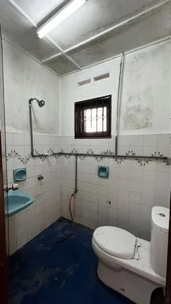 Bathroom features: white ceramic toilet, wall-mounted sink, chrome faucet, blue vinyl flooring, white tiled walls, decorative border tiles, small window with metal bars, exposed plumbing, fluorescent ceiling light.