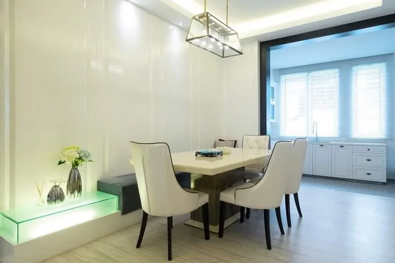 Dining features: rectangular dining table with light wood finish, upholstered dining chairs in cream, chandelier with geometric design, white painted walls, recessed LED lighting, large windows with sheer blinds, built-in cabinetry in white, decorative vases on the table, greenery in decorative pots, wooden flooring; style: modern.