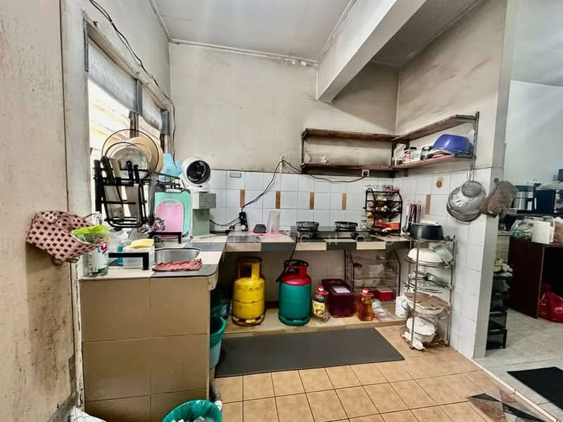 Kitchen features: grey tiled flooring, white painted walls, wooden shelving, stainless-steel sink, gas stove with multiple burners, green gas cylinder, red and green storage containers, kitchen mat, window with sheer curtains, wall-mounted fan.