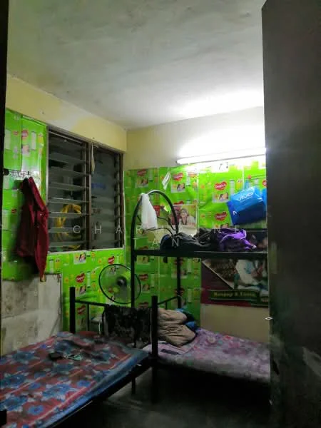Room features: green patterned wallpaper, two single beds with bedding, metal bed frames, window with metal bars, fluorescent ceiling light, concrete flooring, clothes hanging on a rack, various bags and personal items.